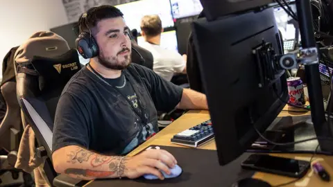 Stephanie Lindgren A young man with heavily tattooed arms sits in front of a computer monitor. His hand rests on a mouse and he wears a large set of over-ear headphones. He looks deep in concentration but calm.