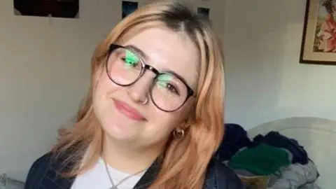 Dakota-Iona/BBC News Delia-Ioana sitting in a room but the crop is too tight to see much of it. She has blonde hair and glasses and is smiling in front of a white wall with framed pictures