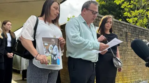 Jo Black/BBC Clare Croucher holding a picture of her daughter. She is standing next to Leah's father John, who is reading a statement to media. Her sister Jade is standing next to her father. 