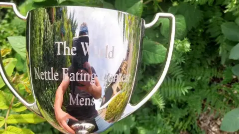 Dorset Nectar Cider A silver-coloured trophy with handles and engraved with the words, the world nettle eating contest men's. A man holding a camera phone can be seen reflected in the metal.