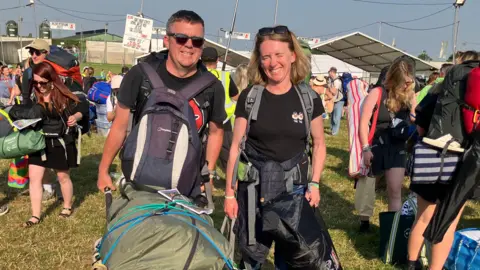 Tom and Emma at the entrance to the site holding backpacks and a large wheelbarrow. 