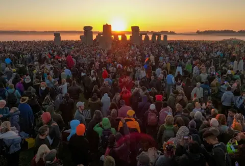 Finnbarr Webster/Getty Images Visitors watch the sunrise at Stonehenge, on June 21, 2024 in Wiltshire, England, 21 June 2024. A crowd stands in the foregorund looking towards the sun which is visible between stones in the background