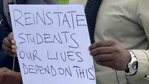 Close up of a man's hand holding a sign saying 'reinstate students our lives depend on this'