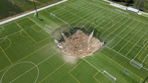 Lawrence Bryant/REUTERS A drone view shows a sinkhole, a large depression of brown soil with pieces of artificial turf hanging from the top edge into the hole, is seen in the middle of a multi purpose sports field at Gordon Moore Park in Alton, Illinois, USA, June 27 2024. 