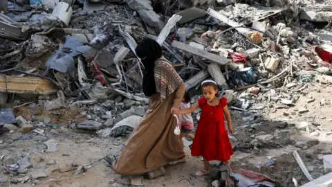 Reuters A Palestinian woman and a girl walk among debris after an operation by Israeli forces to rescue four Israeli hostages who were being held in Nuseirat refugee camp, in the central Gaza Strip (9 June 2024)