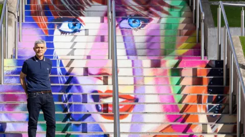 PA Sadiq Khan in a blue T-shirt standing on the steps of the multi-coloured Taylor Swift staircase art