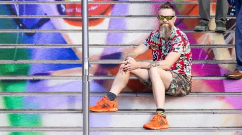 PA Media Frank Styles sitting on the steps, which are covered in a Taylor Swift image. He has a beard and wears sunglasses, and is dressed in shorts and a shirt, with orange trainers