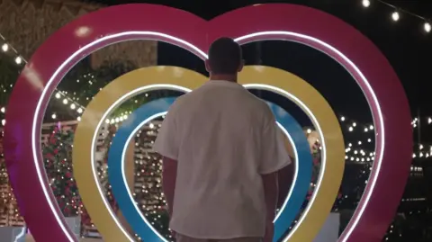 ITV/ Shutterstock Joey Essex entering the Love Island villa. Joey is pictured from the back as he walks through a multi-coloured heart-shaped arch. He wears a white short-sleeved shirt and is pictured outside the villa at night, lit by festoon lighting. 