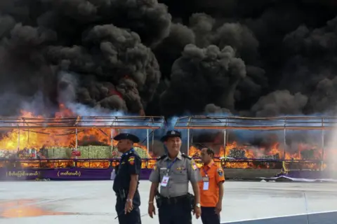 STR/AFP Three officials in uniform stand in front of a burning pile of seized illegal drugs behind a wire fence during a destruction ceremony in Yangon on June 26, 2024.