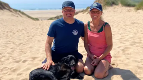 Ollie Bird Ariel, the formerly six-legged dog, lying on a beach alongside her owners who are kneeling