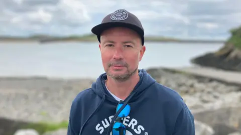 A man wearing a grey cap and a blue hoody with sunglasses hung off the front of the hoody standing by the sea