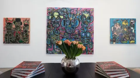 Larkin Durey Three Aboudia paintings hung in a gallery. Copies of his monograph sit on a table in the foreground.