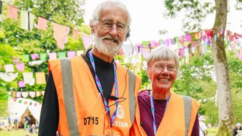 Oxfam  Graham and Judy Cole wearing orange Oxfam hi-vis vests and lanyards. They are standing amongst some trees that have bunting hanging between the branches