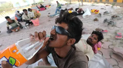 EPA An Indian worker drinks water from a bottle while others rest on the pavement in the shadow of an overpass on a hot day near New Delhi, India, 18 June 2024. 