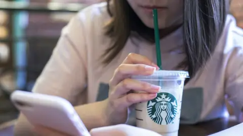 Getty Images Woman sitting at a table drinking cold Starbucks drink through a straw while looking at her mobile phone