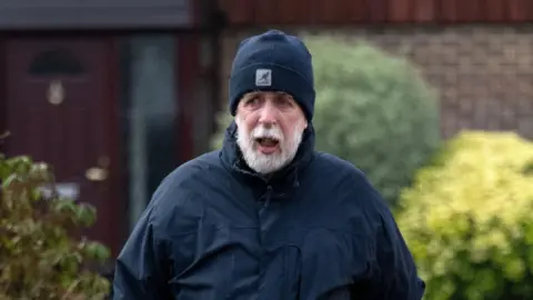 Alamy A grey bearded Gareth Jenkins wearing a dark blue beanie hat and dark blue coat, walking in front of a house