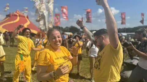 Simon Steele Mr Wilson and Ms Lawrence wearing yellow. They are celebrating and laughing with their hands in the air. Flags and festival tents are visible in the background and there are streamers all over them