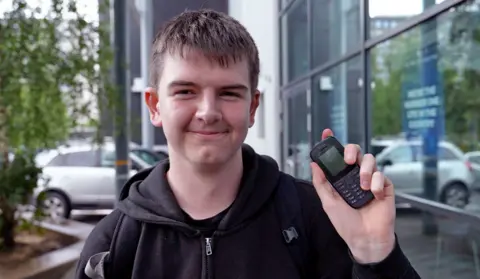 BBC News/Kristian Johnson Will, 15, wears a black hoodie and smiles at the camera as he holds up his basic Nokia phone