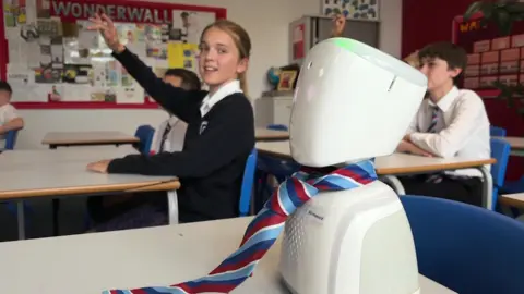 A pupil puts their hand up next to AV Howard, a small white tabletop robot wearing a red and blue striped tie