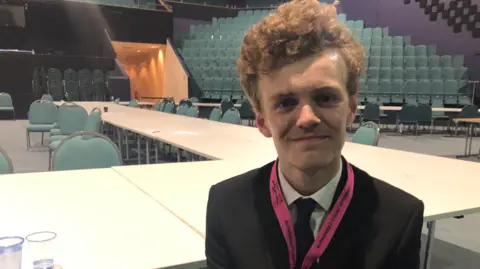 Emma Baugh/BBC Sam Carling, in a jacket and tie, smiling in an empty hall where the election count was held