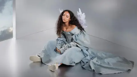 Sony Music Jade Thirlwall in album artwork for her new single. Jade has long curly brown hair worns loose and sits on the floor, her hands in her lap, while wearing a washed denim gown, white trainers and angel wings 