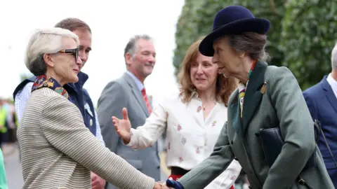 Cameron Smith/PA Wire Princess Anne shaking hands with a woman. Princess Anne is wearing a navy hat, with a green coat. 