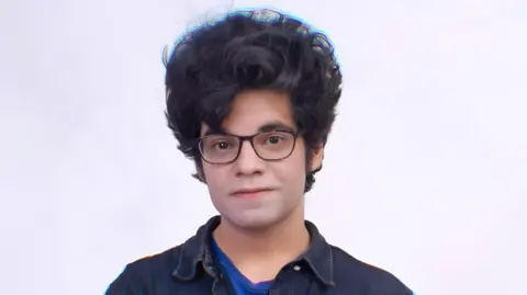 Mano Animations A man with a curly mop of hair and glasses stands in front of a white background. He wears a denim jacket over a dark blue t-shirt.