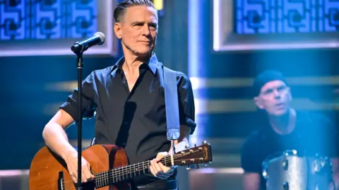 Getty Images Bryan Adams stood in a black shirt playing a guitar, his drummer is behind to the left of him