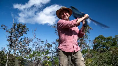 BBC/Tony Jolliffe man in widebrimmed hat swirling a butterfly net with forest and blue sky in background