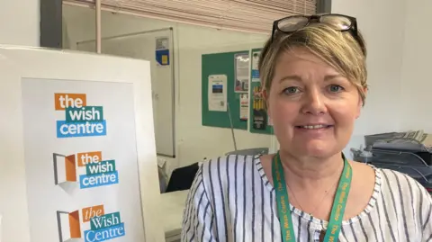 Rebekah Wilson wearing a striped top standing next to a wish centre sign in their office building. Rebekah has blonde hair and glasses on her head and is wearing a green lanyard. 