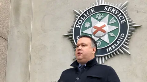 PA Media A man with dark hair in front of a Police Service of Northern Ireland logo