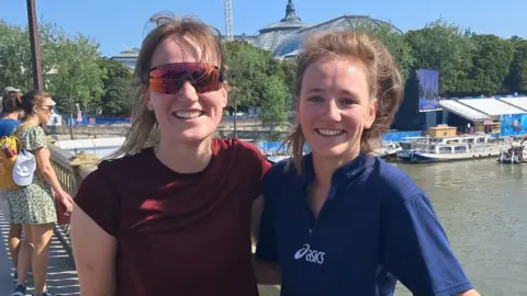 Two women with long dark blonde hair tied up, with their arms around each other - the woman on the left wears a maroon t-shirt, and the woman on the right wears a dark blue t-shirt