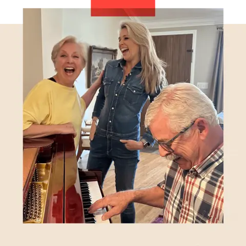 A smiling Marijke stands next to Lara Lewington while Marijke's husband Tom plays the piano