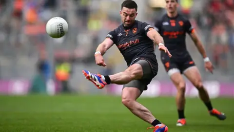 Getty Images GAA footballer in a black kit kicking a white ball during a match. Another man of the same team looks on in the background