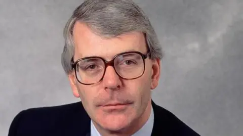 BBC Publicity John Major wearing a suit and brown glasses, smiling directly at the camera