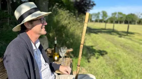 Simon Boas A smiling Simon Boas wearing a panama hat, sitting on a chair beside a green field