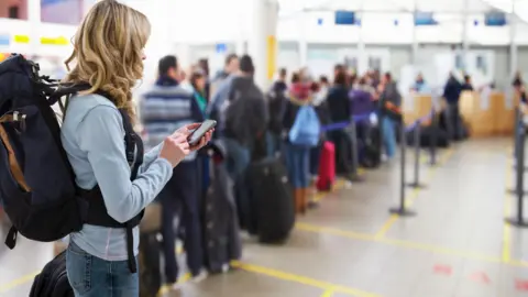 Getty Images Woman carrying backpack and looking at mobile phone while people queue in front of her at the airport