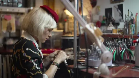 Jewellery designer Jessica De Lotz wearing a berry red beret and a black jacket with drawings of necklaces on, looks down at her hands working. She is sat in front of a desk which is covered with pliers. 