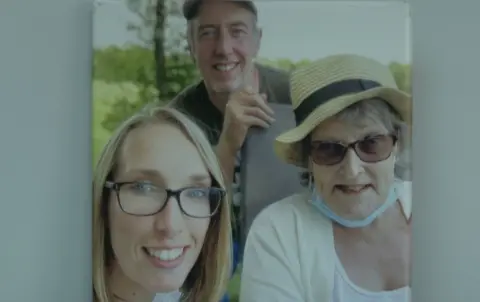 Supplied Anne Clarke wears a straw hat and sunglasses with facemask around her chin. She is with her two adult children.