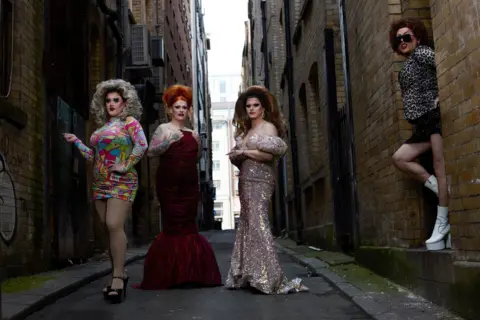 Magnus Hastings Four drag queens pose in an alley way, one wears a rainbow mini dress, another wears a red velvet floor-length gown, the third wears a sparkly gown while the fourth stands in a door way wearing a mini skirt, a leopard print blouse and white go go boots