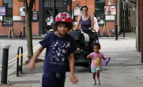 Thea walking down a London street with Isaac in a pram, JJ walking ahead of her and Moses on a scooter in the foreground