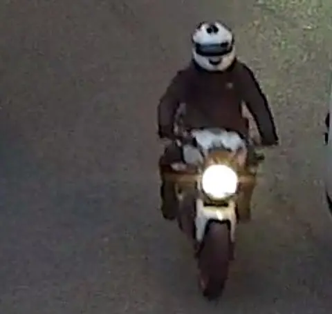 Met Police CCTV image of the motorcyclist from a front view showing him in black on a white motorbike with a big circle headlamp.