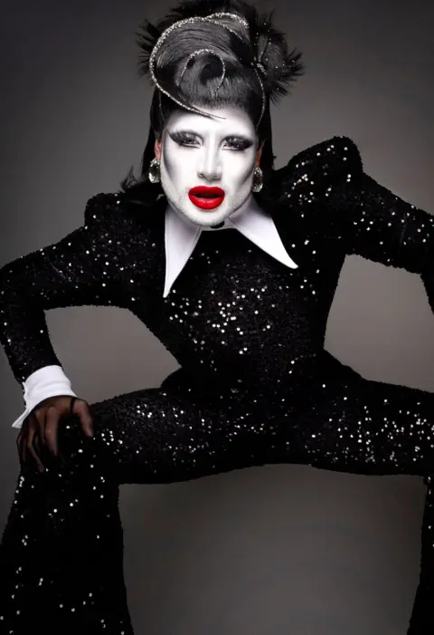 Mangus Hastings A drag queen with a white face and a sparkly black jump suit poses for the camera, wearing bright red lip stick and a black feathered wig.