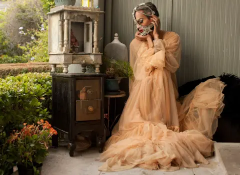 Magnus Hastings A drag queen in a flowing tuille dress sits next to ruined antique furniture. She has a diamond and pearl encrusted mask around her mouth and draped over her head