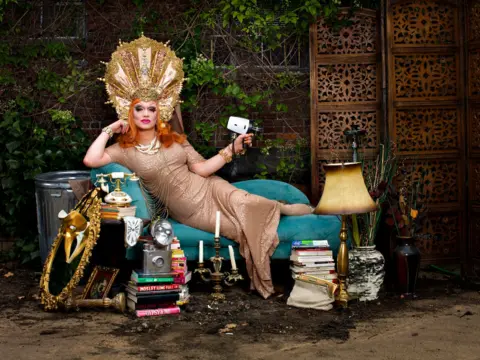 Magnus Hastings A drag queen with red shoulder-length hair drapes herself over a chaise lounge while wearing a gold headdress. She is surrounding by antique artefacts and is holding an old vintage camera