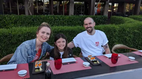 Emma Williams-Tully Emma, sitting in a restaurant with plates of sushi on the table, with her husband Dan and their nine-year-old daughter in the middle