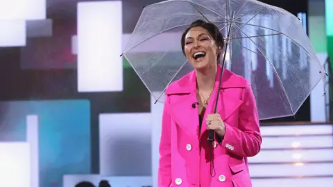 Getty Images Emma presenting Big Brother in 2018, she is wearing a pink coat and holding a clear umbrella