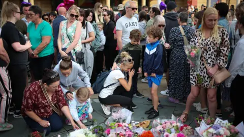 EPA A crowd of people, ranging from young children to grey-haired adults, light candles and leave floral tributes to the victims of the Southport attack, some bowing their heads in sadness, others holding each other for comfort