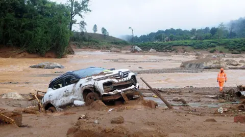 Reuters A rescuer walks past a damaged car at a landslide site after multiple landslides in the hills in Wayanad