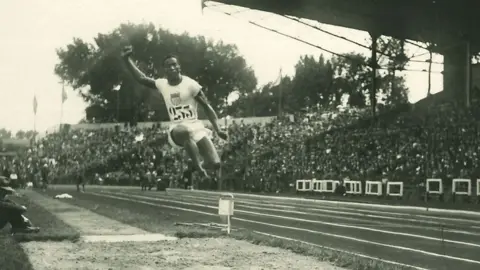 Collections Musée National du Sport, France A close-up of a black and white postcard showing William DeHart Hubbard in mid-air doing a long jump in 1924